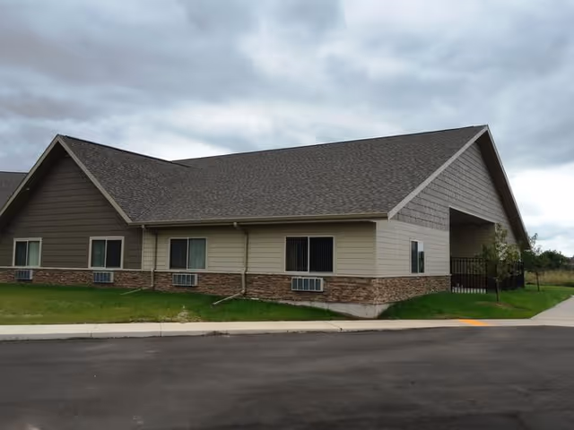 Exterior view of a single-story senior living facility building with a large pitched roof, windows, stone siding, and a small lawn under a cloudy sky.