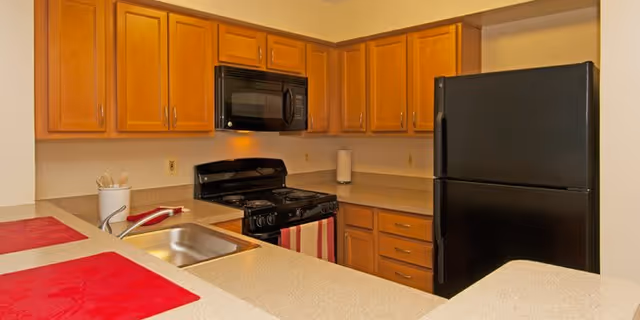 Small kitchen with honey-colored cabinets, black refrigerator, stove and microwave, a sink and countertops with red placemats.