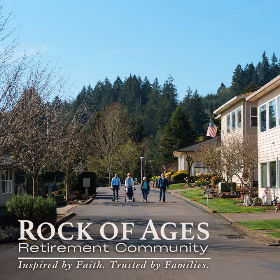 Exterior view of Rock of Ages Retirement Community with residents walking along a street beside beige buildings and trees.