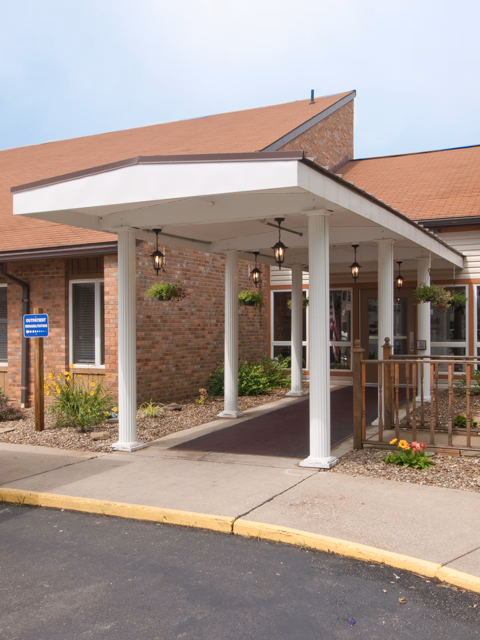 Covered entrance with white columns, hanging lanterns, and landscaping at the front of a brick senior living building.