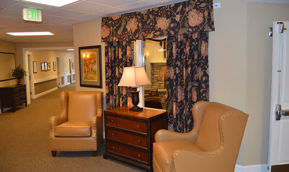 A cozy seating area in a hallway of Walnut Creek Memory Care facility featuring two tan leather armchairs positioned on either side of a wooden chest of drawers with a table lamp on top. Behind the chest is a window with floral-patterned curtains. The hallway extends into the background with framed pictures on the walls and an exit sign overhead.