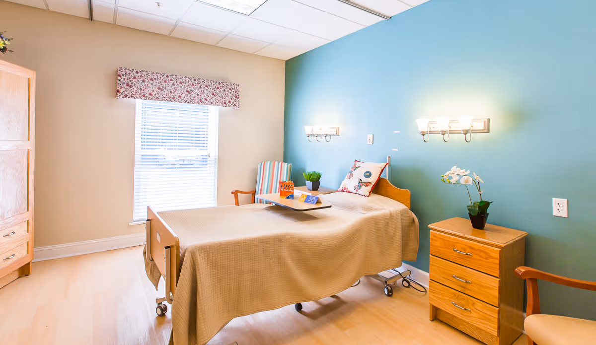 A bright and clean nursing home bedroom with a single bed covered in a beige blanket. There is a wooden nightstand with a potted orchid, a striped armchair, and a wooden wardrobe. The walls are painted beige and teal, and there is a window with blinds and a floral valance letting in natural light.