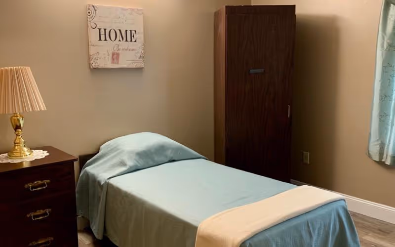 A small bedroom with a single bed covered in light blue bedding and a cream-colored throw at the foot. Next to the bed is a dark wooden nightstand with a gold lamp and a doily. On the wall above the bed is a decorative sign that reads 'HOME'. A dark wooden wardrobe stands in the corner near a window with light blue curtains.