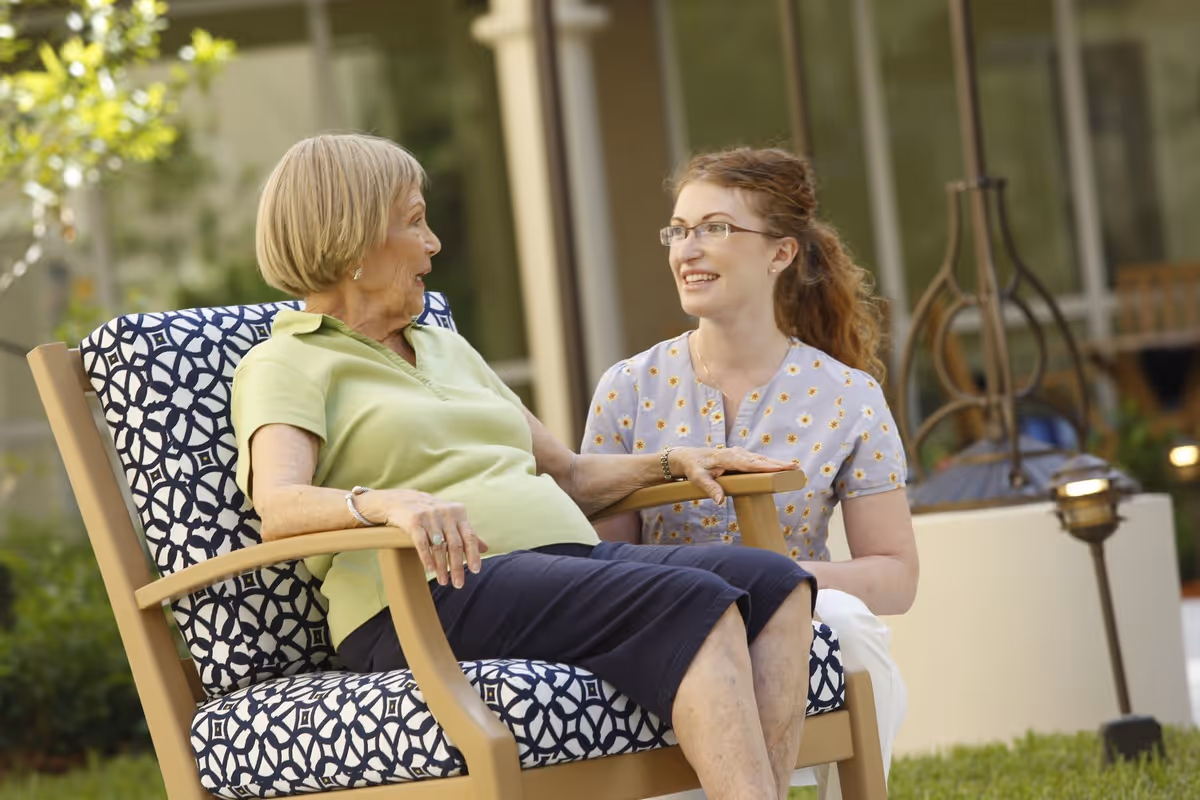 An elderly woman sitting on a patterned cushioned outdoor chair, engaged in a conversation with a younger woman kneeling beside her. They are outside in a garden or patio area with greenery and a building visible in the background.