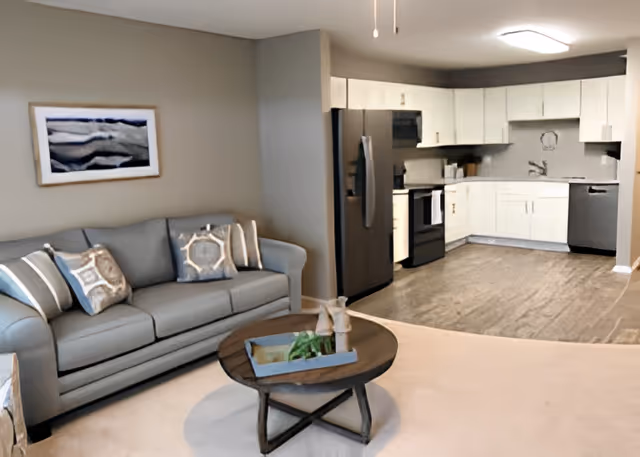 A modern open-concept living area and kitchen in a senior living facility. The living area features a gray sofa with patterned cushions and a round wooden coffee table with decorative items. The kitchen has white cabinets, stainless steel appliances including a refrigerator, stove, and dishwasher, and a light wood floor.