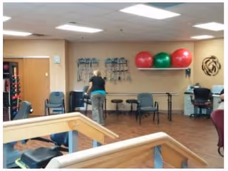 Interior view of a senior living facility exercise or therapy room with wooden flooring, exercise balls on a wall shelf, chairs, tables, and a person standing near the back wall. Various exercise equipment is visible, and the room is well-lit with ceiling lights.