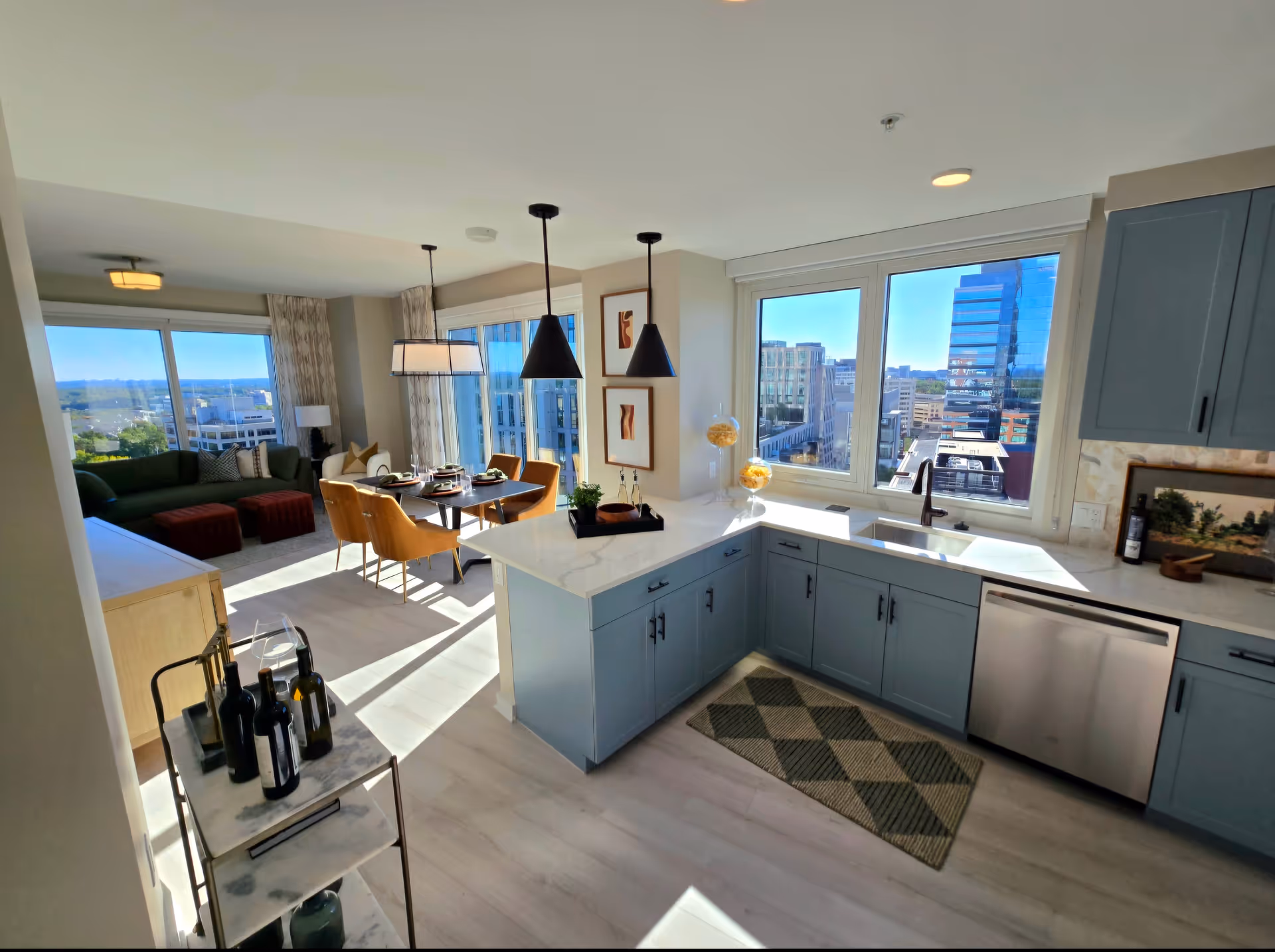 Bright modern kitchen and dining area with large windows showing a cityscape view. The kitchen has blue cabinets, a white countertop, a sink under the window, and a dishwasher. The dining area has a table set for four with mustard yellow chairs and pendant lights hanging above. A living room with a green sofa and ottomans is visible in the background.