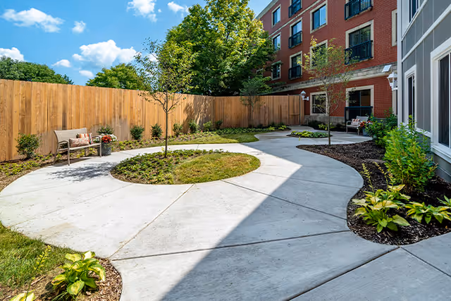 Sunlit enclosed courtyard with a circular concrete walkway, planted beds, benches, and the adjacent brick building.
