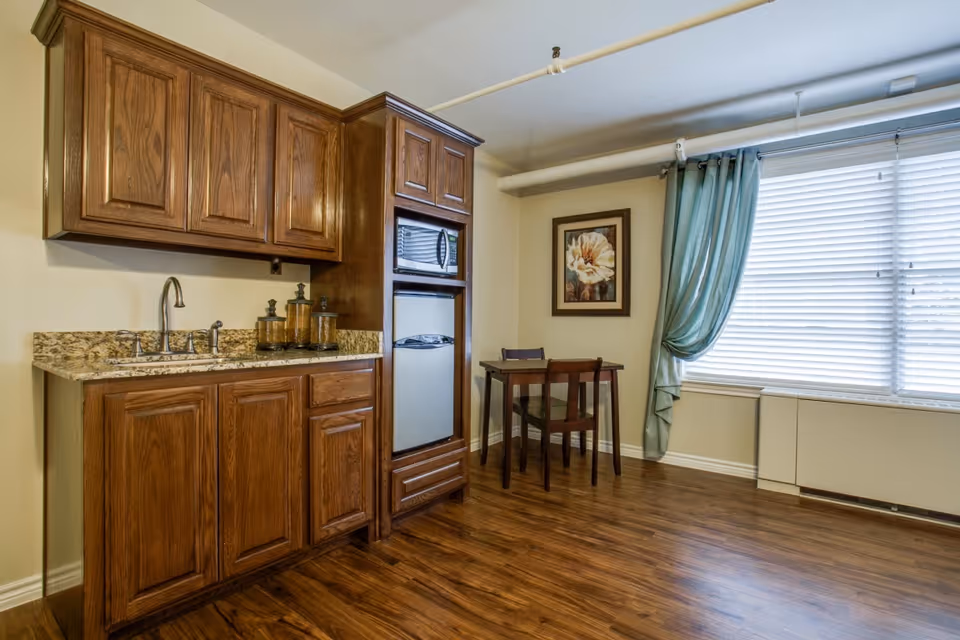Small kitchenette with wooden cabinets, a sink, microwave and mini-fridge, and a small dining table by a window with teal curtains.