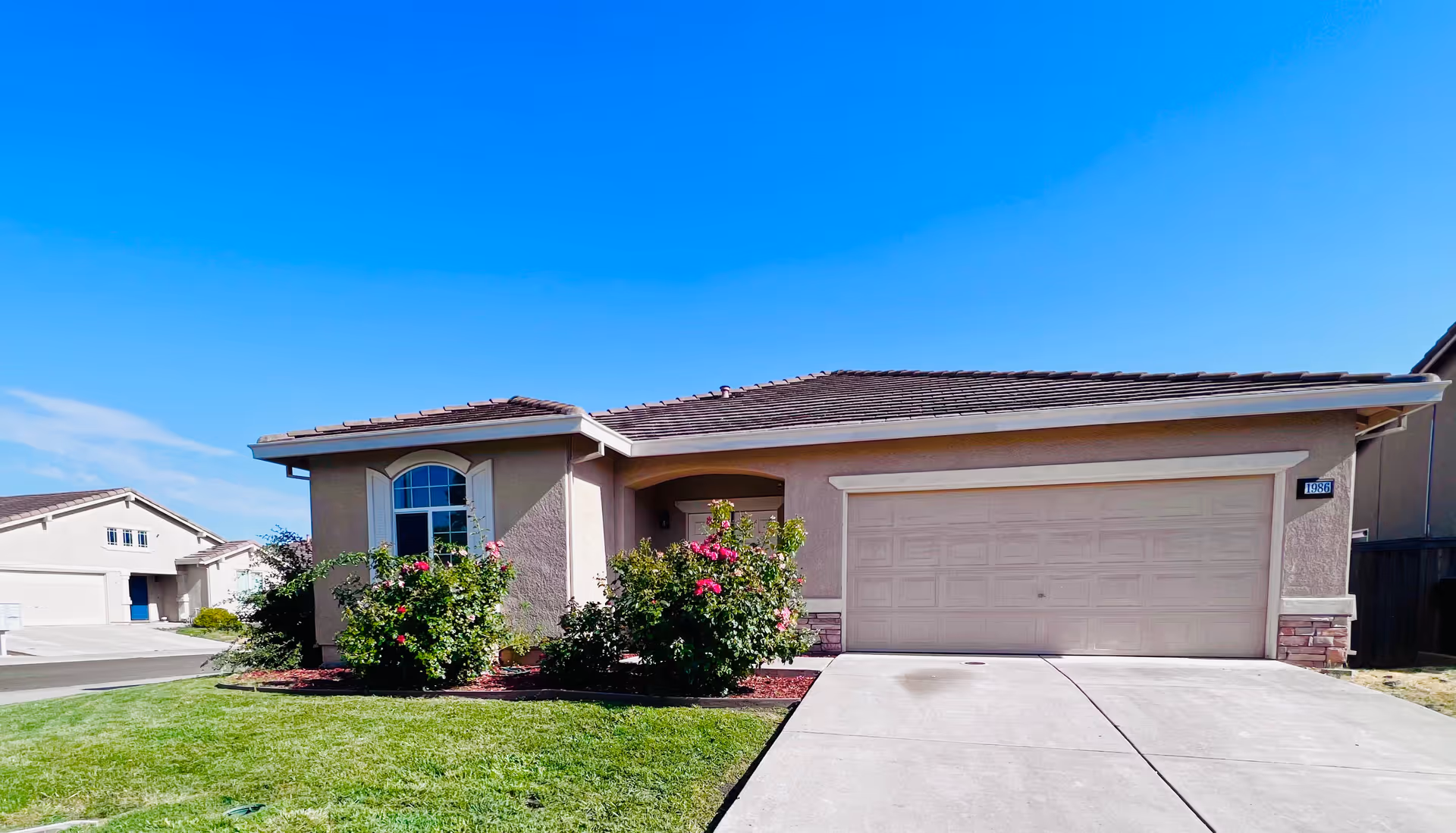 Single-story suburban house with a two-car garage, beige exterior walls, a tiled roof, and a small front garden with green grass and flowering bushes under a clear blue sky.