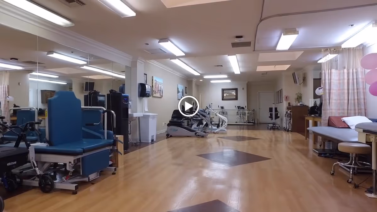 A spacious rehabilitation room with wooden flooring and exercise equipment including stationary bikes and a wheelchair. The room has bright overhead lighting, a large mirror on one wall, and medical examination tables with stools on the right side. There are curtains and some balloons in the corner, suggesting a welcoming environment.