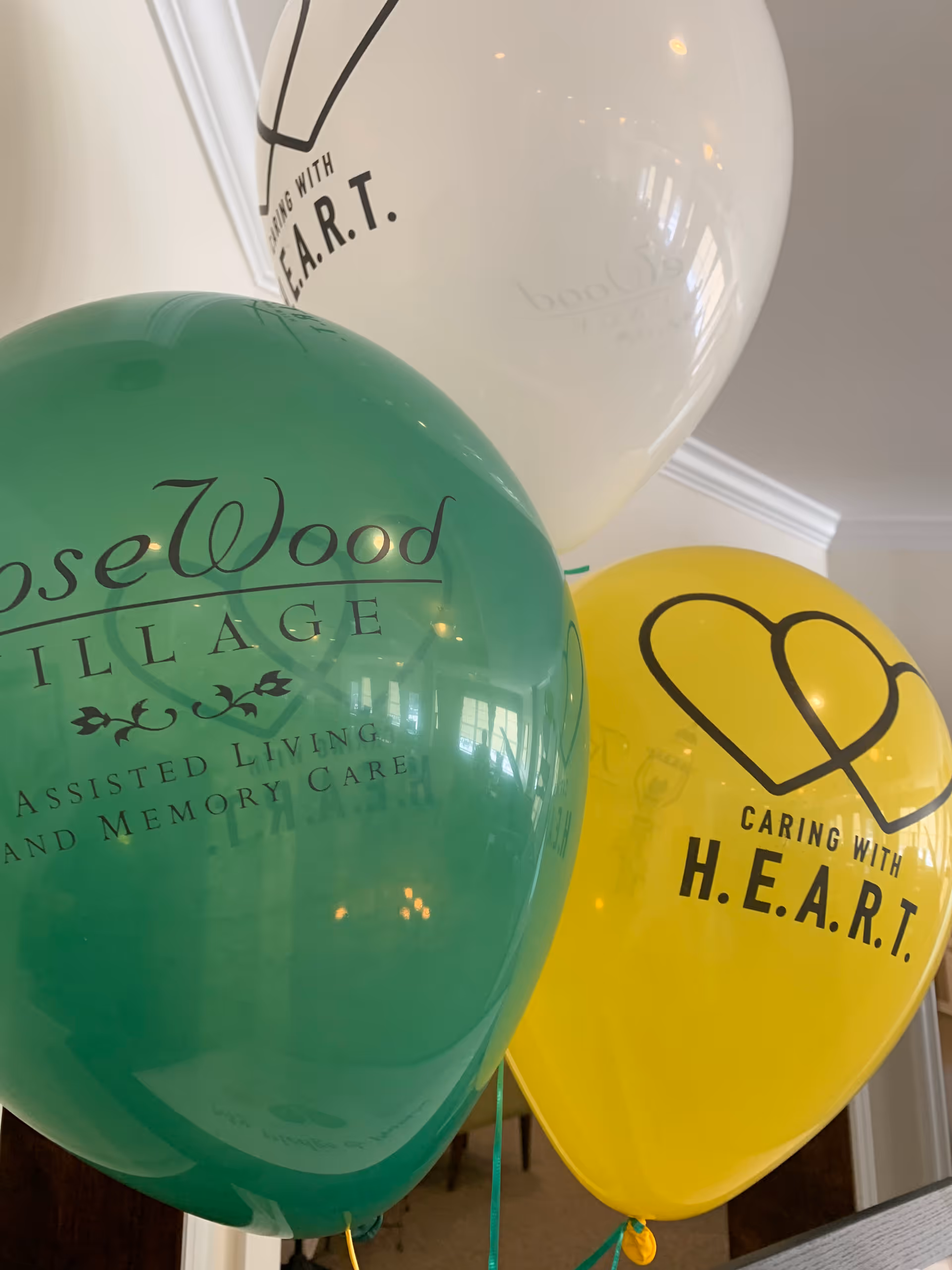 Three balloons indoors with printed text. A green balloon reads 'RoseWood Village Assisted Living and Memory Care.' A yellow balloon has a heart graphic and the text 'Caring with H.E.A.R.T.' A white balloon also has the text 'Caring with H.E.A.R.T.' The background shows part of a room with white crown molding and a window reflection.