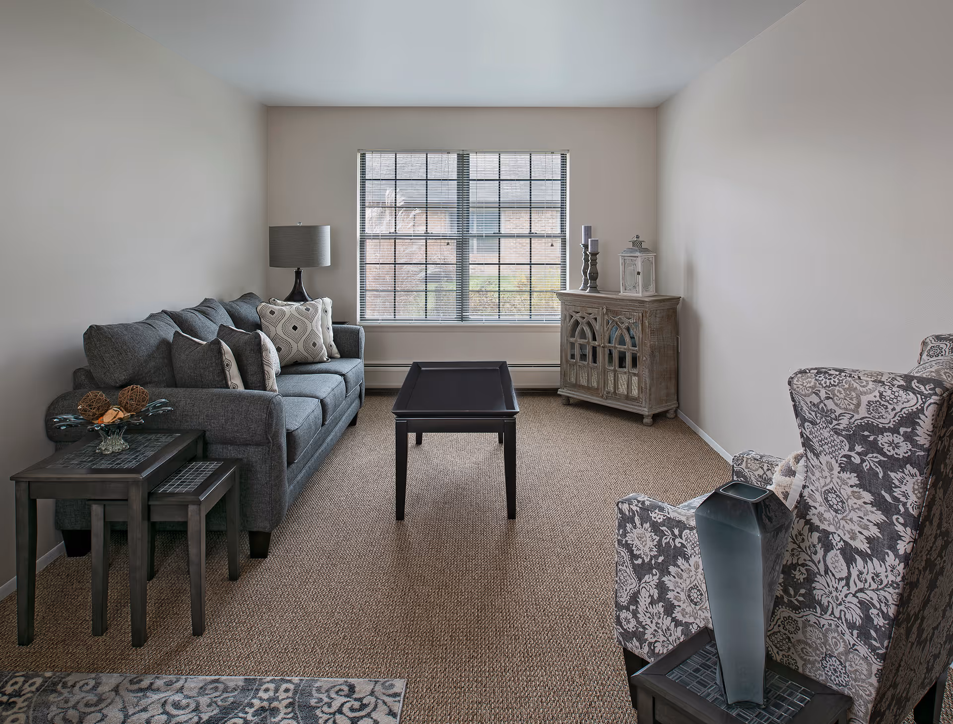 A cozy living room with a gray sofa adorned with patterned cushions, a black coffee table in the center, a wooden side cabinet with decorative items near a large window with blinds, and a patterned armchair with a vase on a small side table.