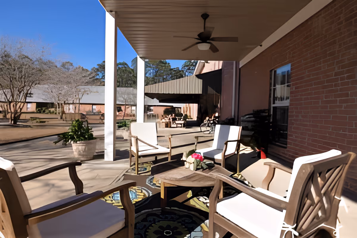 Outdoor covered patio area with four cushioned wooden chairs arranged around a wooden coffee table with a small flower arrangement. The patio overlooks a courtyard with trees and additional seating areas, under a clear blue sky.
