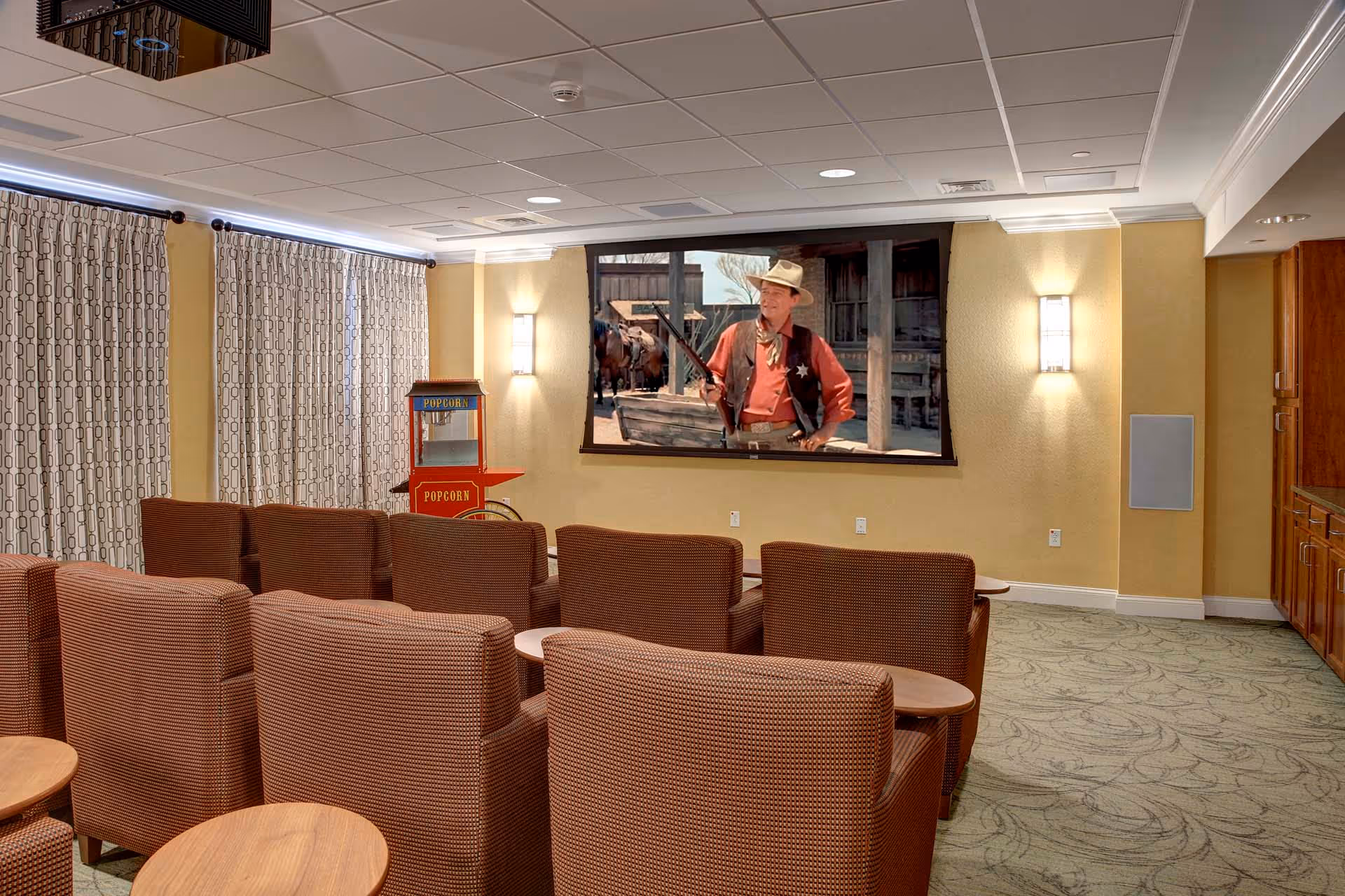 A small theater room with rows of brown upholstered chairs facing a large screen showing a western movie scene. There is a popcorn machine in the corner near patterned curtains, and the room has soft yellow walls with wall sconces providing light.