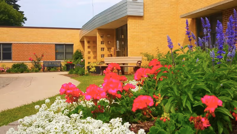 Exterior view of a yellow brick building with a garden in the foreground featuring vibrant red, white, and purple flowers. A concrete pathway curves alongside the garden leading to the building entrance, which has a bench nearby.
