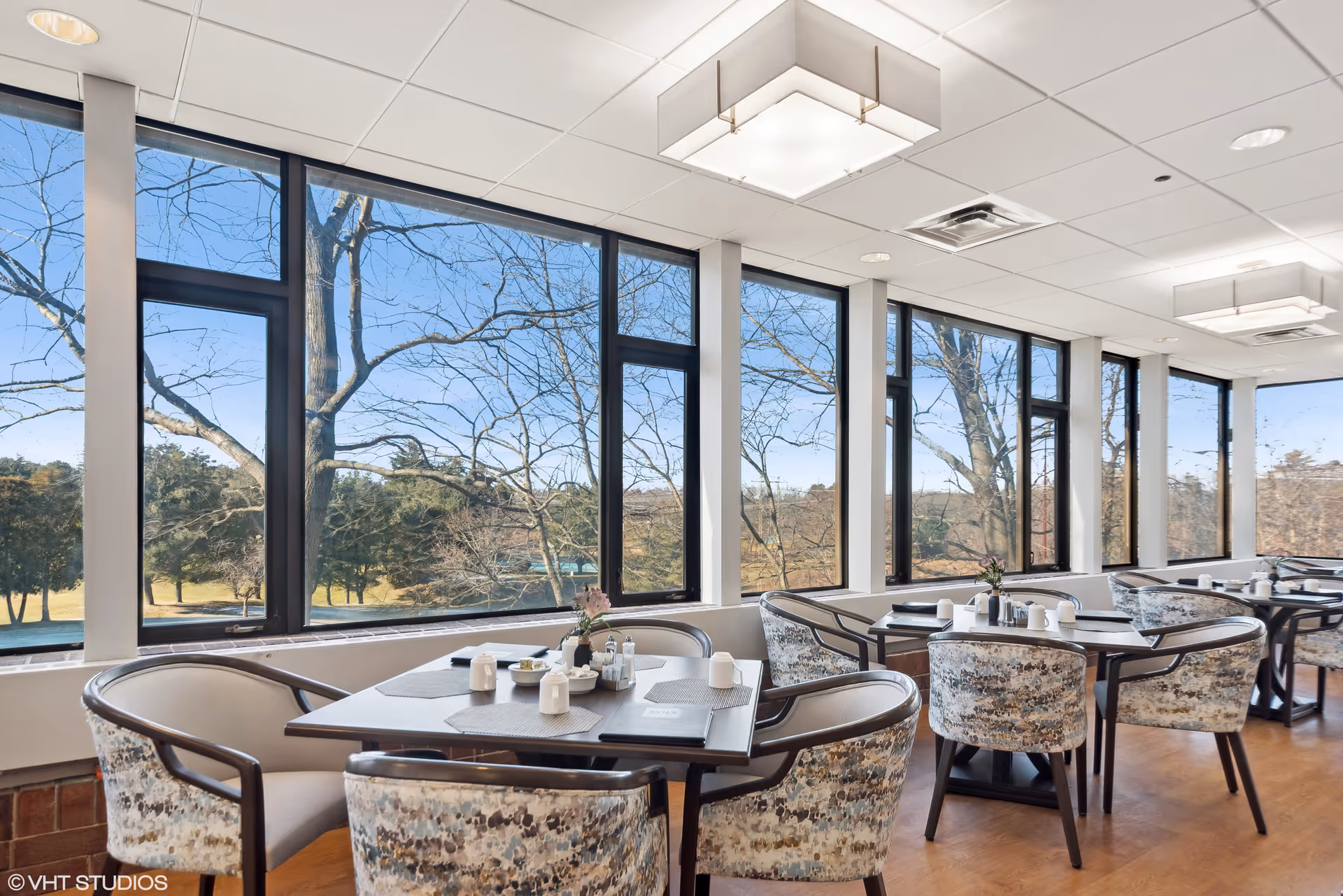 A bright dining room with large windows offering a view of trees and a clear blue sky. The room features several tables with patterned chairs, each table set with placemats, menus, and small condiment containers. Modern ceiling lights illuminate the space.