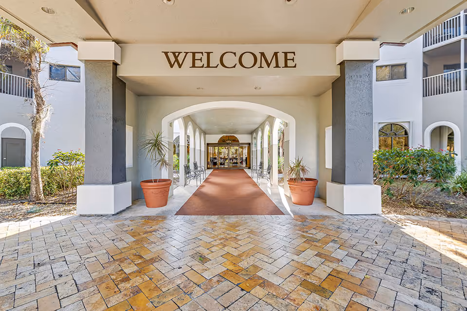 Entrance area of a senior living facility with a covered walkway featuring a 'WELCOME' sign overhead, potted plants on either side, benches along the walls, and glass doors at the far end leading inside the building.