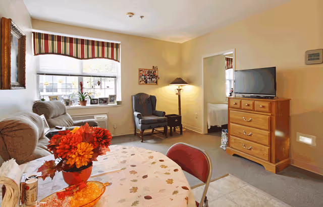 A cozy living area in a senior living facility with a round table covered with a floral tablecloth and a vase of red and orange flowers. There is a beige recliner chair near a window with striped valance, a blue upholstered armchair next to a floor lamp, and a wooden dresser with a flat-screen TV on top. An open doorway leads to a bedroom with a bed visible. The walls are painted light beige and the room is softly lit.