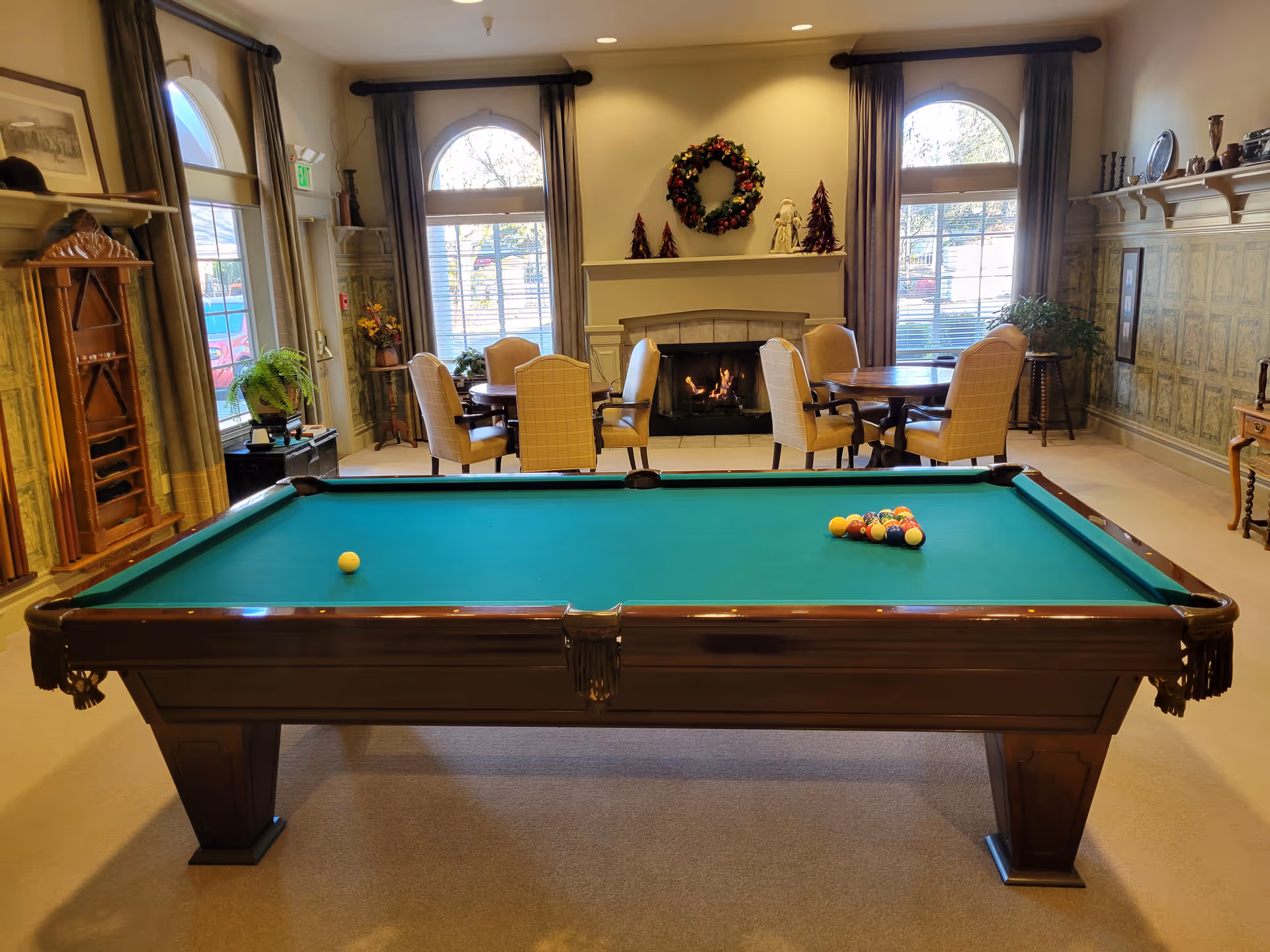 A billiards table in a community lounge with seating, a lit fireplace, and large arched windows.
