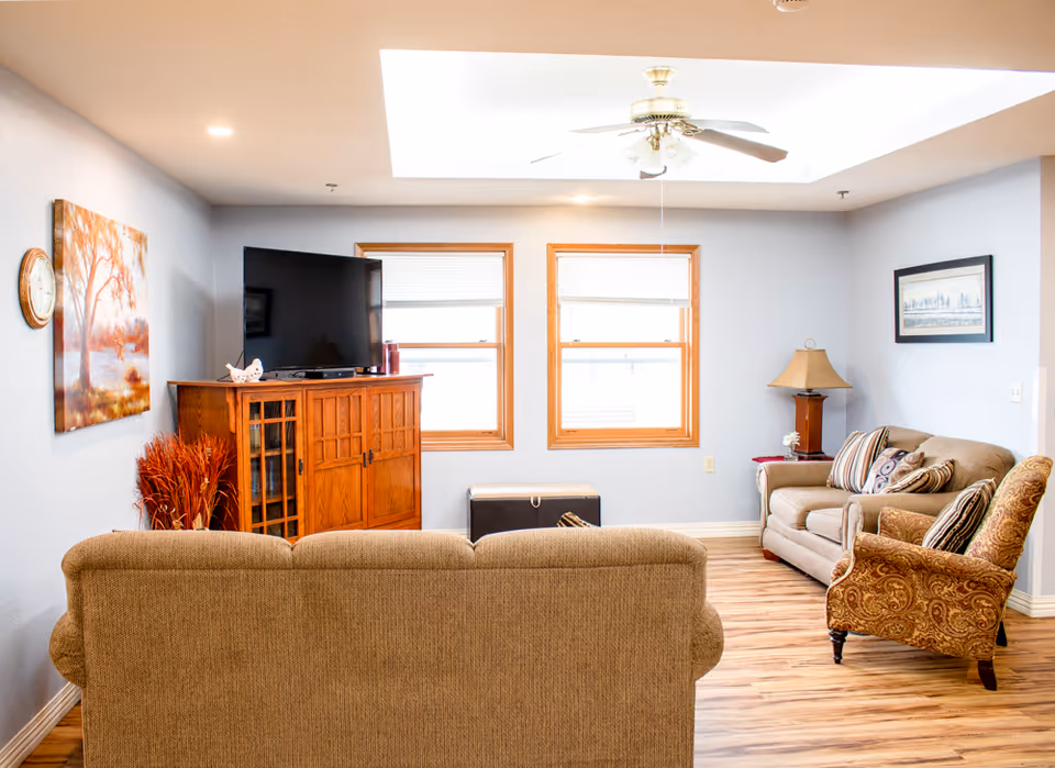 A cozy living room with light blue walls and wooden flooring. The room features a beige sofa facing away from the camera, a wooden cabinet with a flat-screen TV on top, two windows with blinds, a beige loveseat with patterned cushions, a patterned armchair, a wooden side table with a lamp, and framed artwork on the walls. A ceiling fan with lights is mounted on the ceiling.