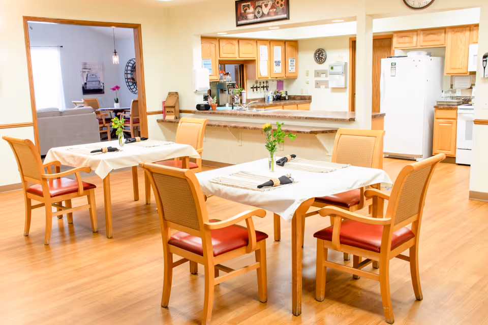 Communal dining room with set tables and wooden chairs in front of a serving counter and open kitchen.
