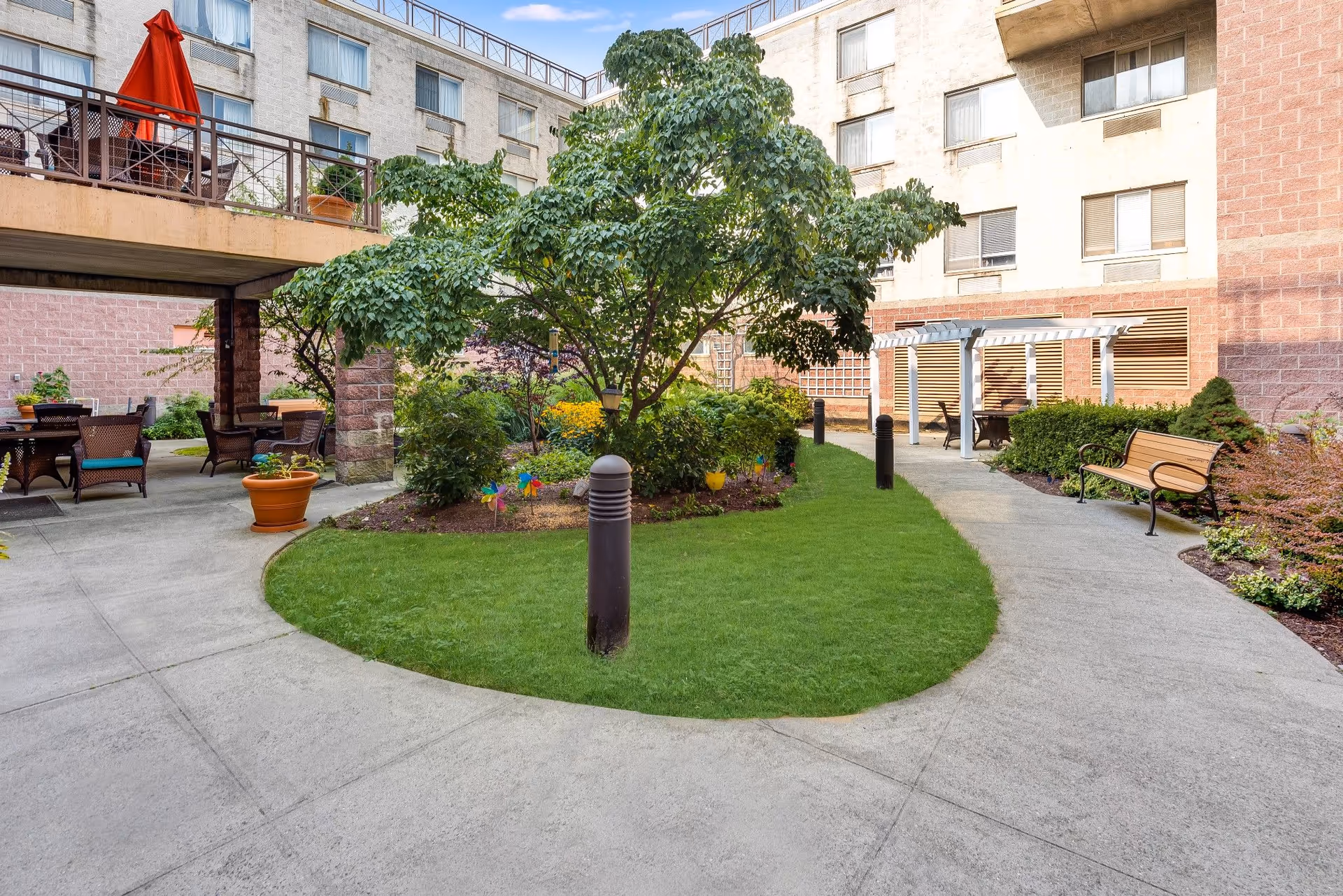 Outdoor courtyard area of a senior living facility with a central tree surrounded by green grass and flower beds. There are paved walkways curving around the garden, outdoor seating with tables and chairs, a bench, and a white pergola structure. The building with multiple windows surrounds the courtyard.
