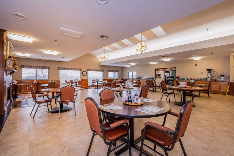 A spacious dining room in a senior living facility with multiple round tables and chairs arranged neatly. The room has large windows with valances, a high ceiling with chandeliers, and a buffet area with coffee machines and food service equipment along one wall.