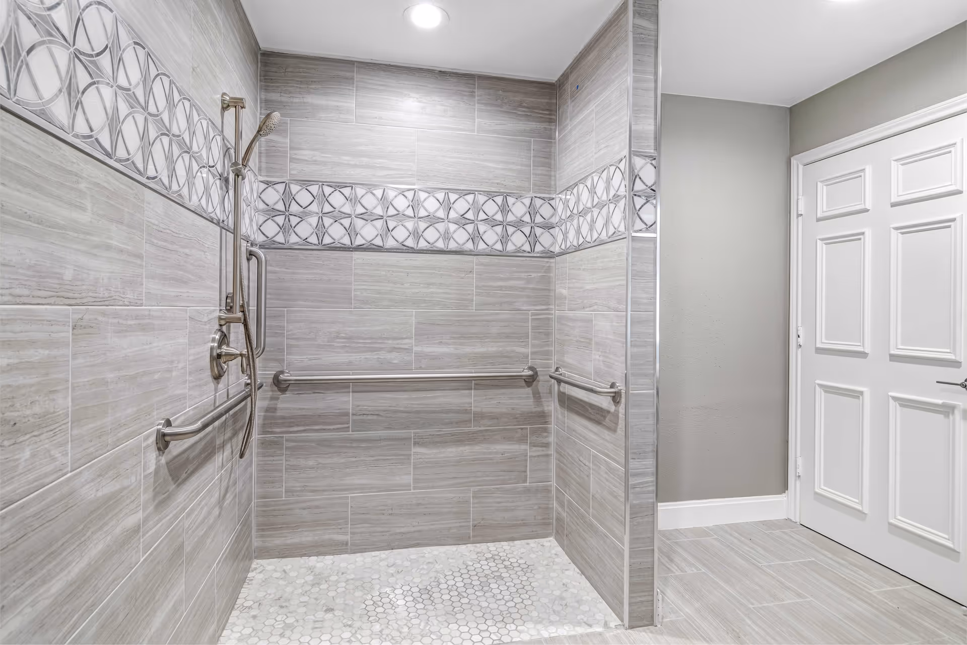 A modern, accessible shower area with light gray tiles on the walls and floor. The shower features a handheld showerhead, multiple grab bars for safety, and a decorative tile border with circular patterns. A white door and gray painted wall are visible outside the shower area.