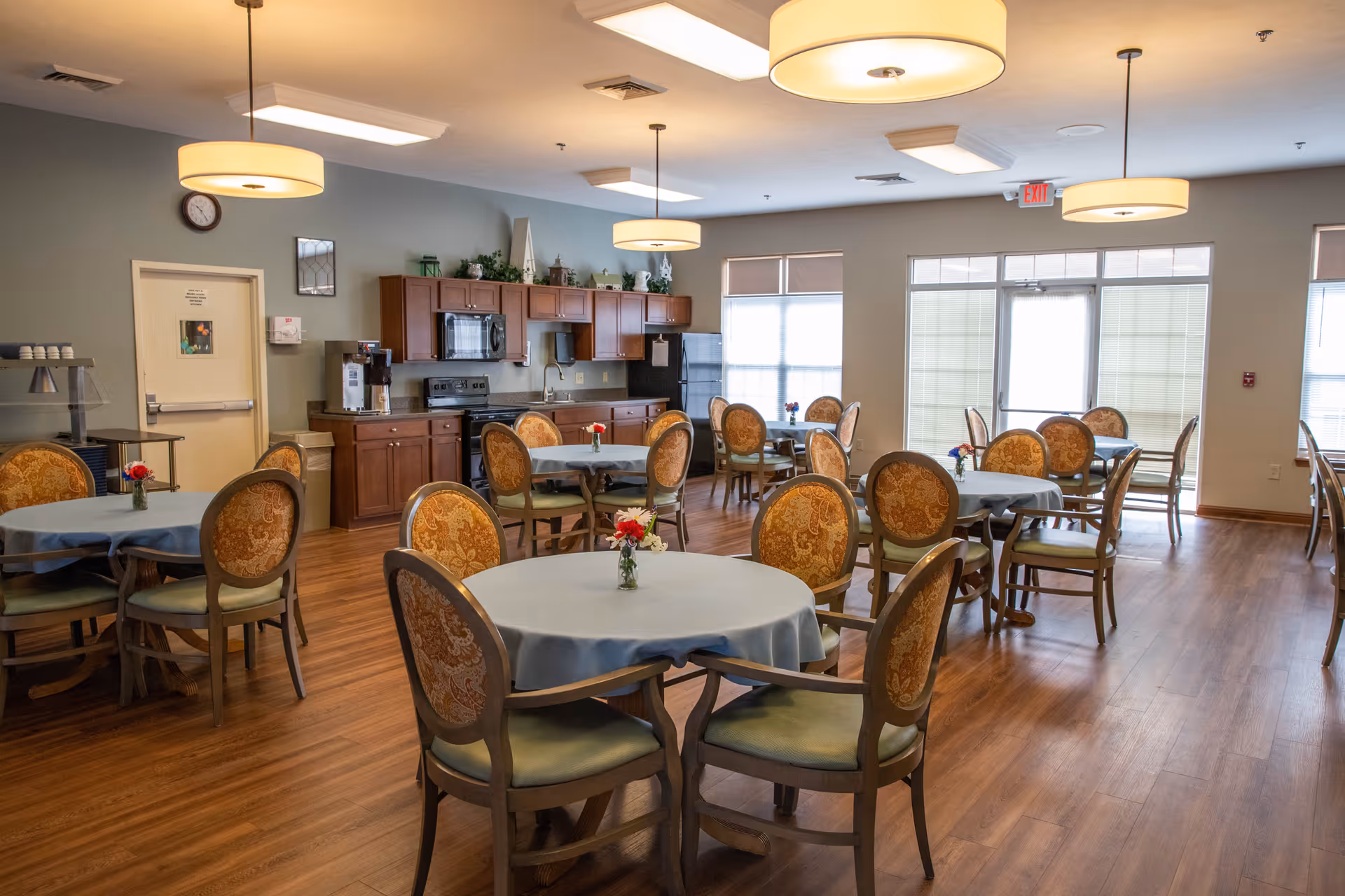 A dining room in Meadow Lakes Assisted Living and Garden Homes featuring multiple round tables covered with light blue tablecloths, each surrounded by ornate wooden chairs with patterned upholstery. The room has wooden flooring, large windows with blinds, and a kitchen area with wooden cabinets, a microwave, and a stove in the background. Ceiling lights illuminate the space warmly.