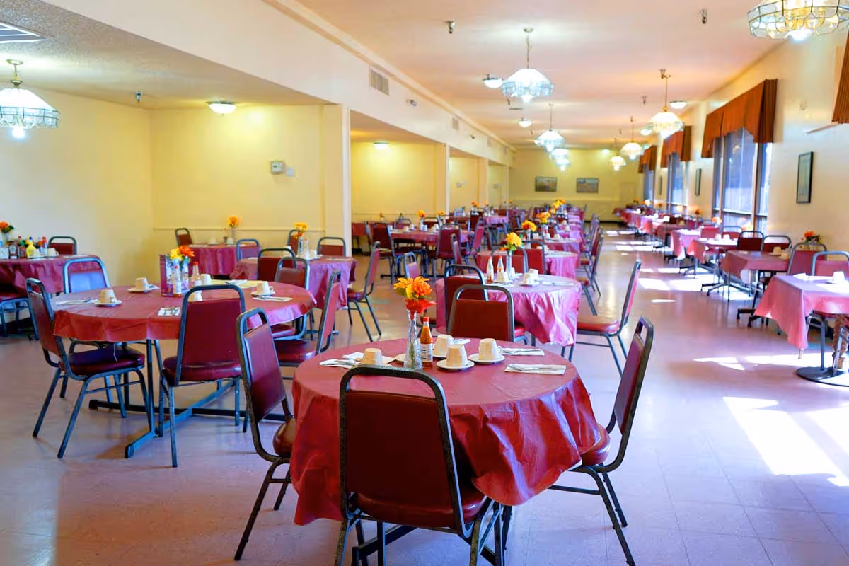 A large dining room with multiple round tables covered in red tablecloths, each set with white cups, saucers, and utensils. Chairs with red cushions surround the tables. The room is well-lit with ceiling lights and natural light coming through large windows with red valances. Small flower arrangements are placed on each table.
