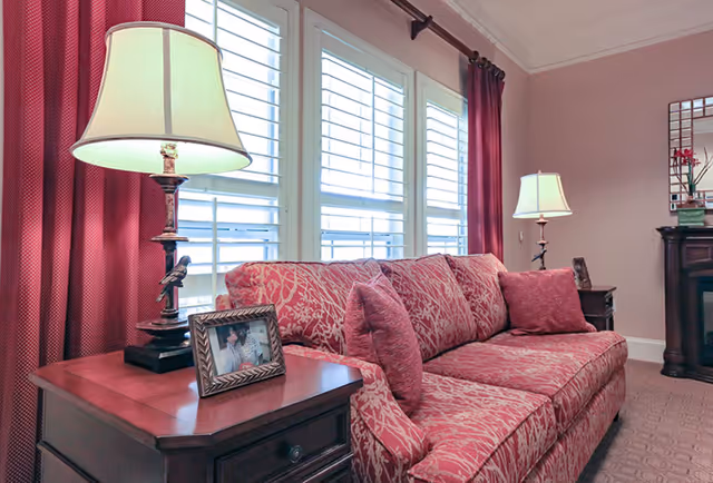 A cozy living room area with a patterned red sofa and matching cushions. There are two wooden side tables, each with a lamp featuring a beige lampshade. One side table also holds a framed photo. Behind the sofa are large windows with white shutters and red curtains. The walls are painted light pink, and a decorative mirror and fireplace are visible in the background.