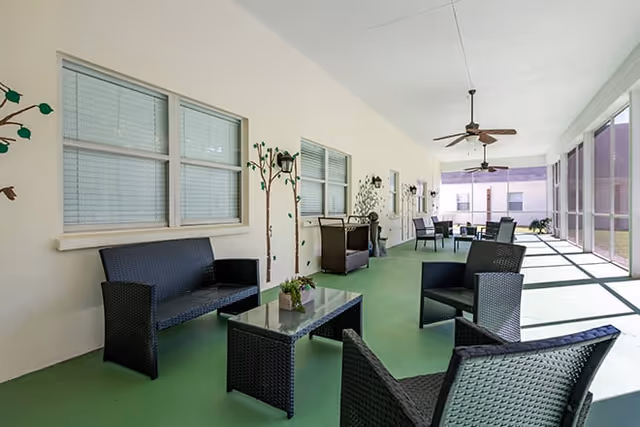 A spacious screened-in porch area with green flooring, black wicker seating including chairs and a sofa, glass-top tables, ceiling fans, and decorative wall art featuring tree designs. The porch overlooks an outdoor area with sunlight streaming in.