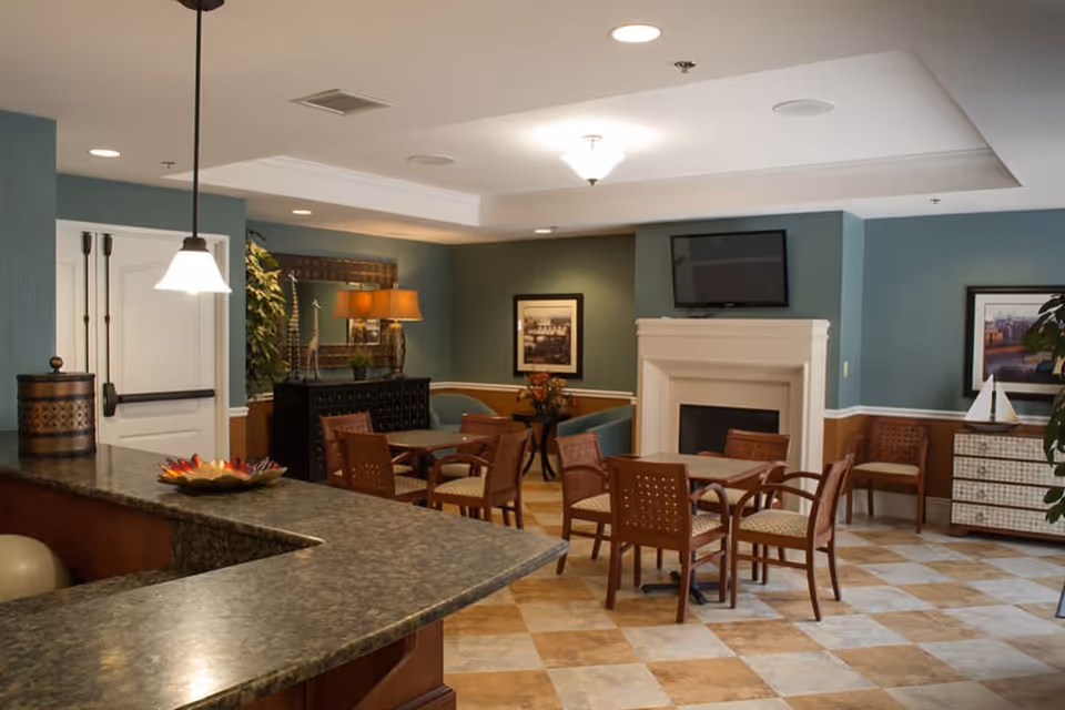 Community dining and lounge area with a granite counter in the foreground, multiple wooden tables and chairs, a fireplace with a wall-mounted TV, and ambient lighting.