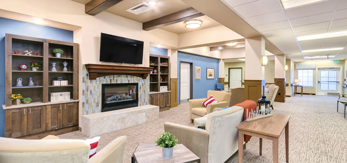 A cozy senior living facility common area featuring a fireplace with a mounted flat-screen TV above it, flanked by wooden shelves with decorative items. The room has beige and blue walls, carpeted floors, and comfortable armchairs with red and white striped pillows. A wooden table with a 'welcome' sign and a small plant is in the foreground. The space is well-lit with ceiling lights and wall sconces, and a hallway with doors and windows is visible in the background.