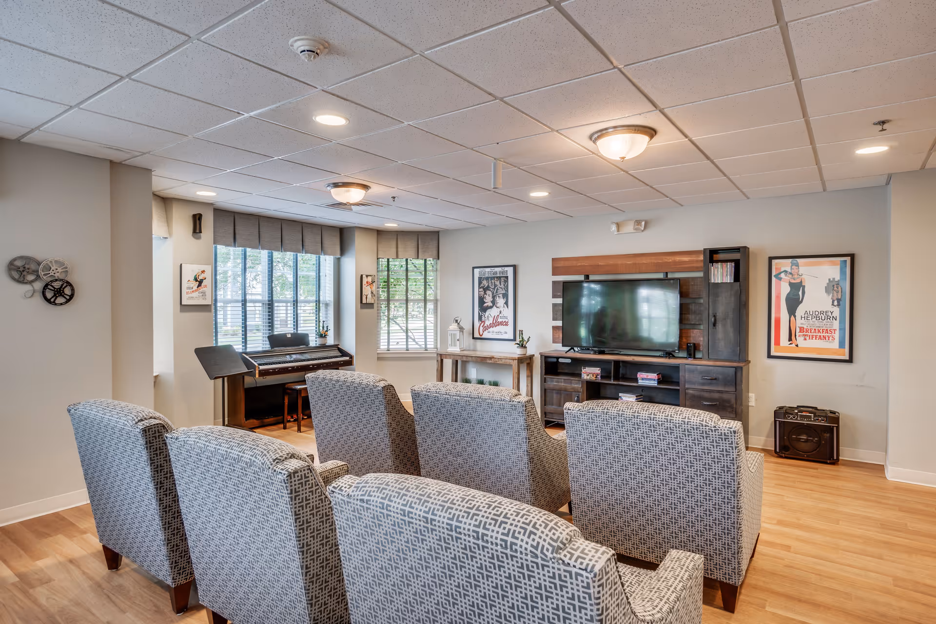 A cozy living room area with six patterned armchairs arranged in two rows facing a large flat-screen TV mounted on a wooden entertainment center. The room has light-colored walls, wood flooring, and a ceiling with recessed lighting and two ceiling fixtures. There are two large windows with blinds and valances letting in natural light. A piano is positioned near the windows, and framed vintage movie posters decorate the walls.