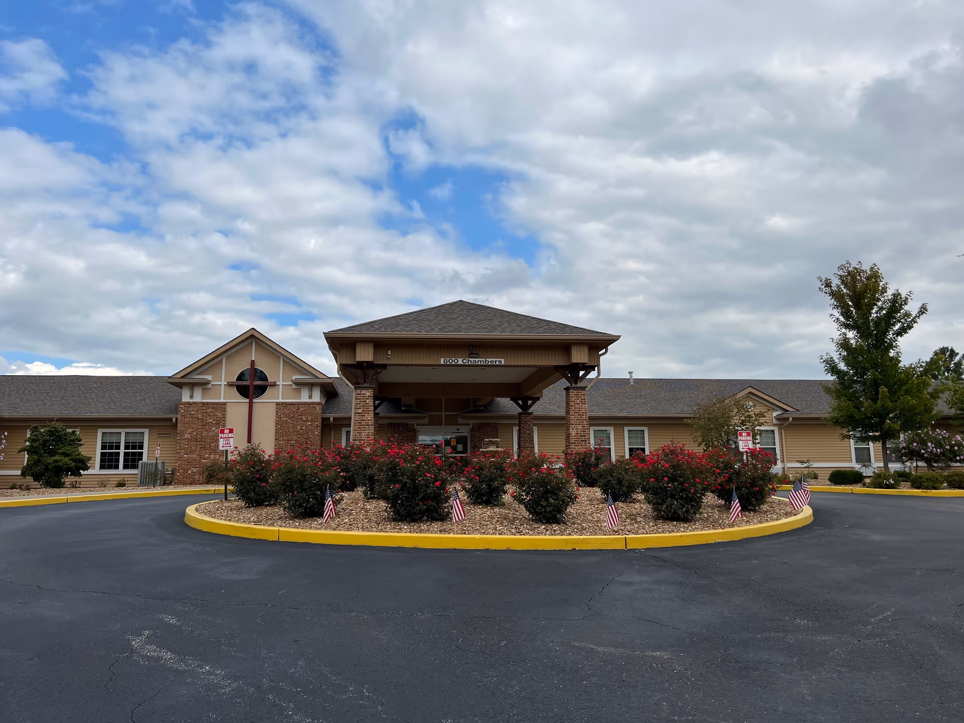 Front exterior view of Arbor Hills Nursing and Rehabilitation Center showing a single-story building with a covered entrance, brick and beige siding, a circular driveway with landscaped bushes and small American flags, under a partly cloudy sky.