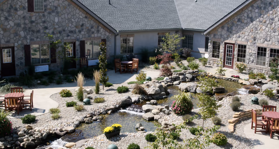 Outdoor courtyard area of a senior living facility with stone buildings surrounding a landscaped garden featuring a small pond with rocks and waterfalls, various plants, and multiple wooden tables with chairs on concrete patios.