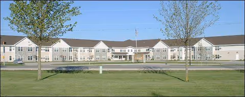 Front view of a long two-story beige senior living building with a manicured lawn, two trees, and an American flag.