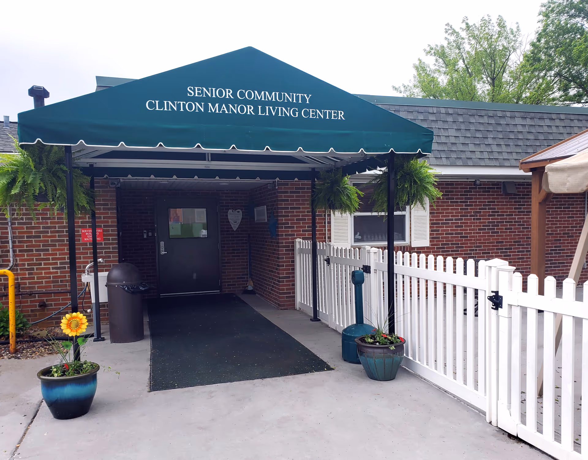 Entrance to Clinton Manor Living Center senior community with a green canopy displaying the facility name. The entrance has a black door, brick walls, white picket fence, potted plants, and hanging ferns.