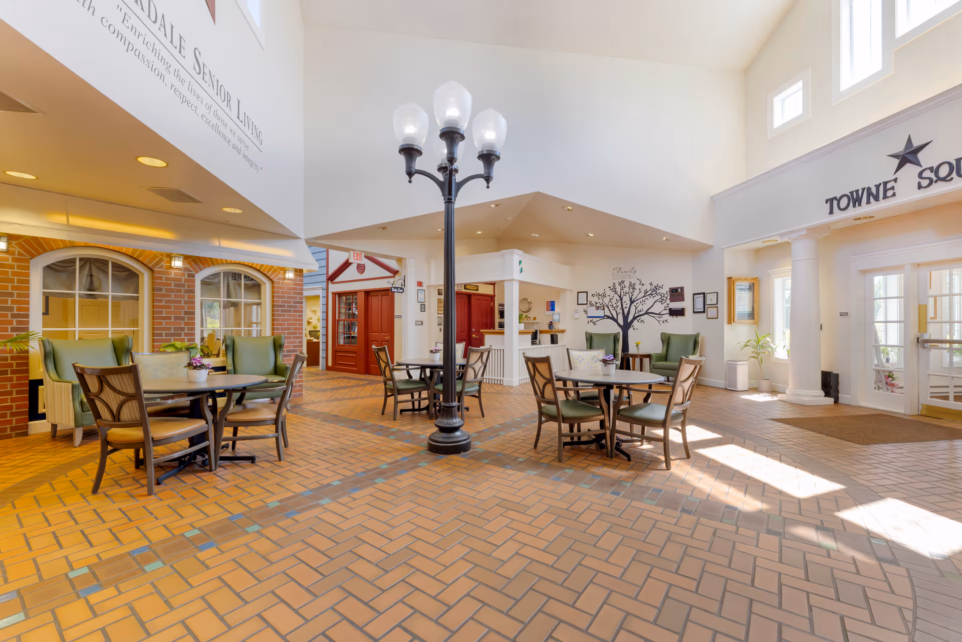 A bright and spacious common area in a senior living facility with brick-patterned tile flooring, several round tables with chairs and green armchairs, a decorative black streetlamp-style light fixture in the center, and large windows letting in natural light. The walls feature a family tree decal and the words 'Towne Square'.