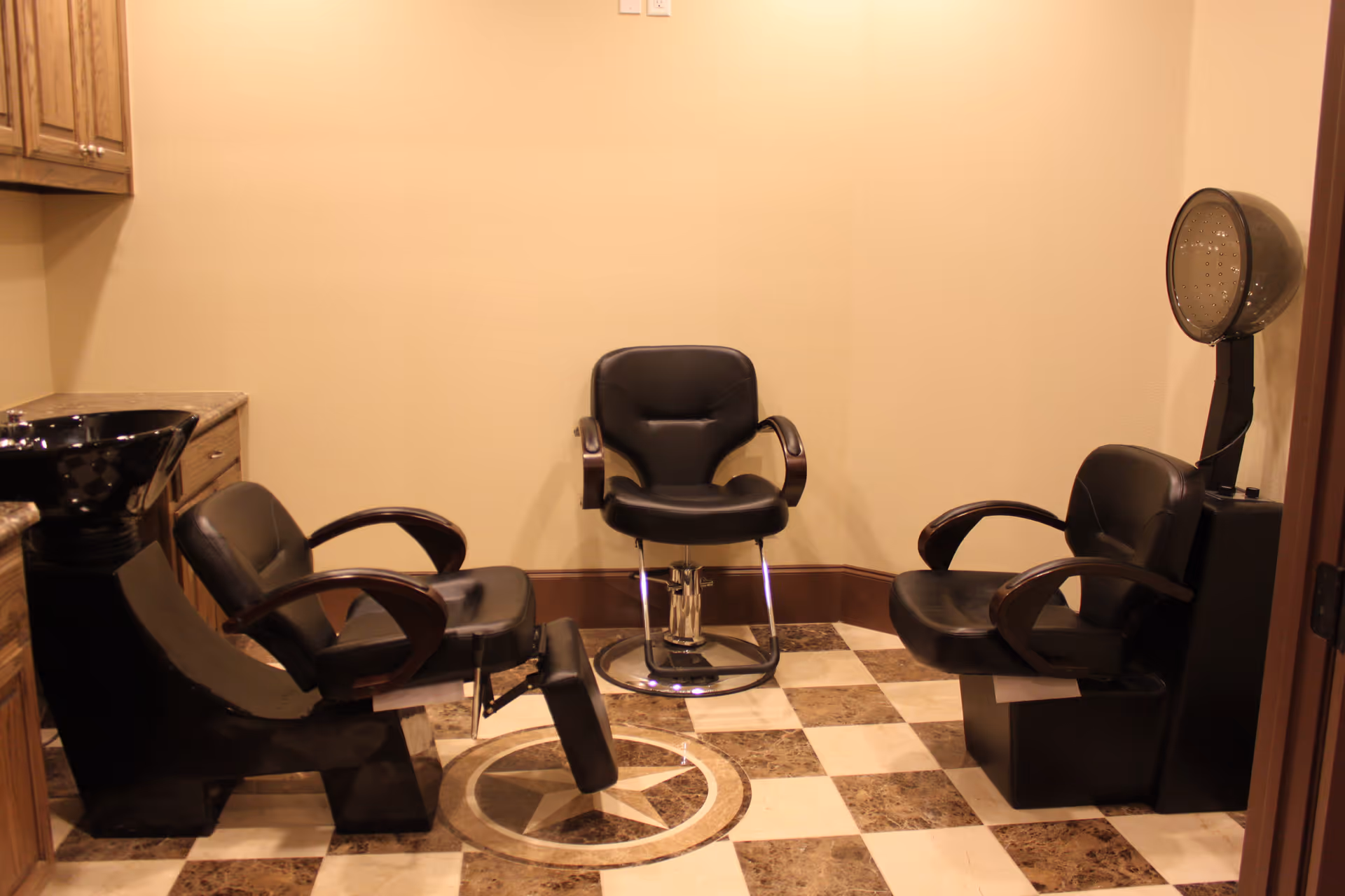 Interior view of a salon area with three black salon chairs, one with a footrest, a black hair washing sink, wooden cabinets, and a hair dryer hood. The floor has a checkered pattern with a star design in the center.