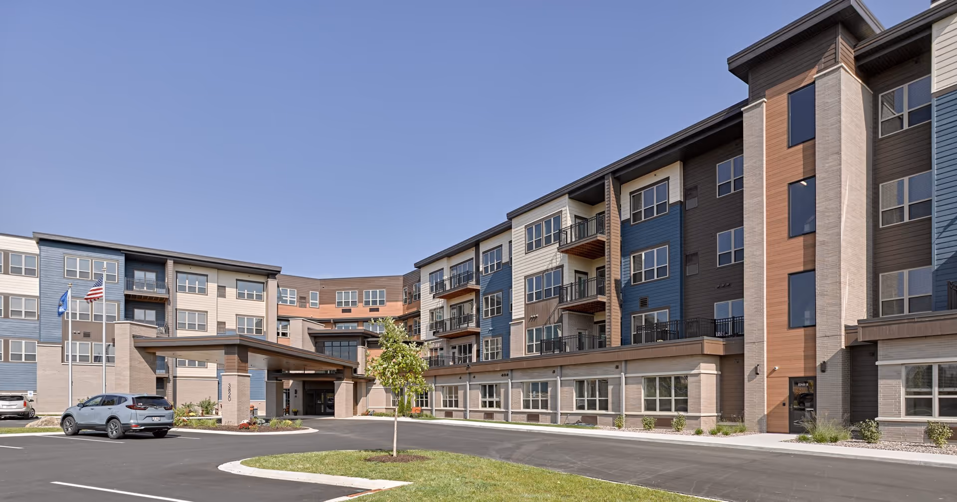 Exterior view of Cardinal View Senior Living facility showing a modern multi-story building with balconies, large windows, a covered entrance, a parking lot with cars, and a clear blue sky.