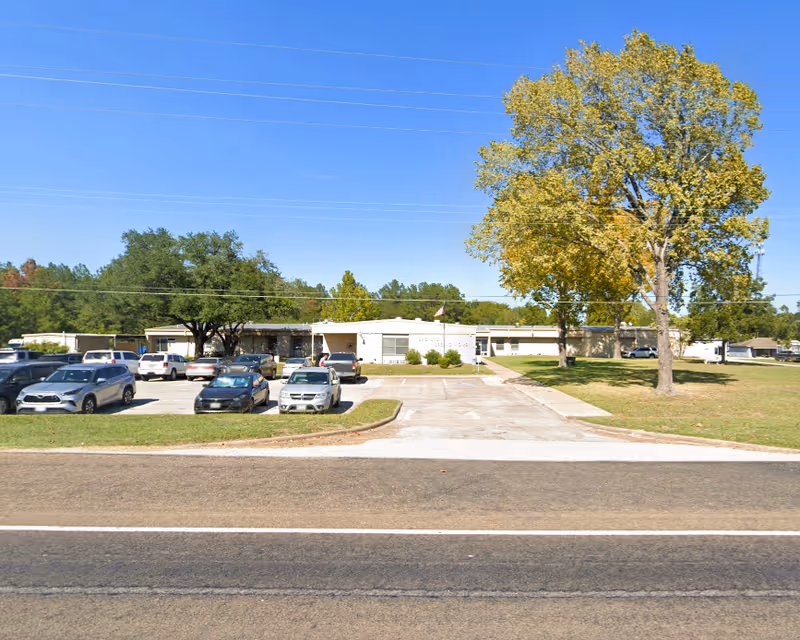 Front view of a single-story nursing home building with a parking lot, several cars, and large trees under a clear blue sky.