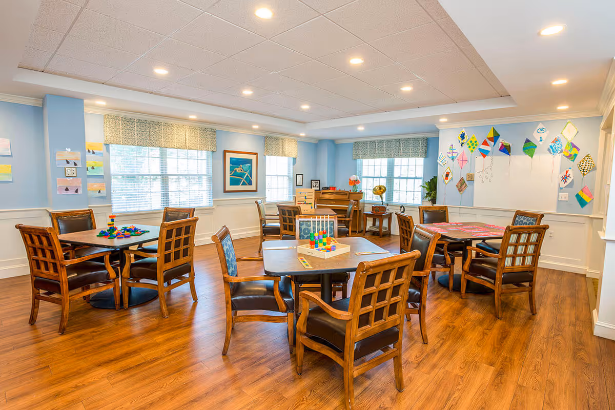 A bright and spacious activity room with wooden floors and light blue walls. The room contains multiple square tables surrounded by wooden chairs with leather cushions. Various colorful toys and games are placed on the tables. Large windows with patterned valances allow natural light to fill the room. The walls are decorated with framed artwork and colorful kite drawings. A piano and a gramophone are positioned near the windows in the background.