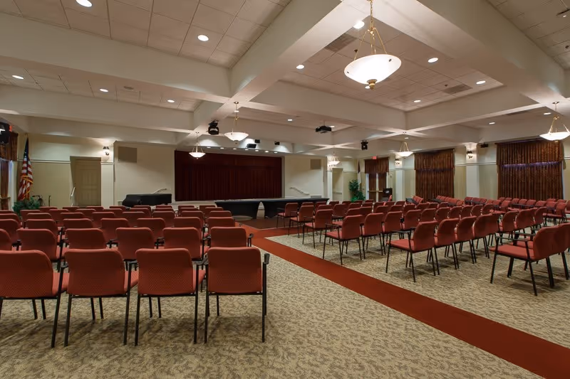 A large room set up with rows of red cushioned chairs facing a stage with a closed curtain. The room has beige walls, carpeted floors with a red runner, and several hanging light fixtures. An American flag is visible on the left side near a door.