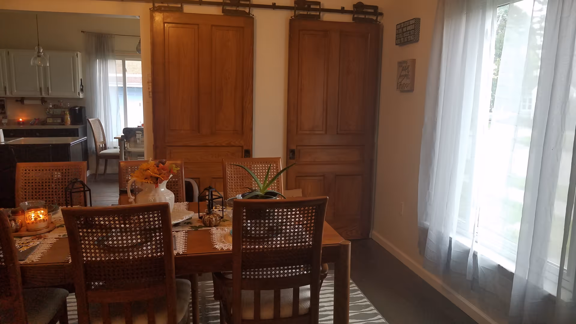 A cozy dining room with a wooden table set with decorative items including a vase with flowers, candles, and a plant. There are six wooden chairs around the table. The room has two wooden sliding doors and a large window with sheer white curtains letting in natural light. A kitchen area is visible through an open doorway in the background.