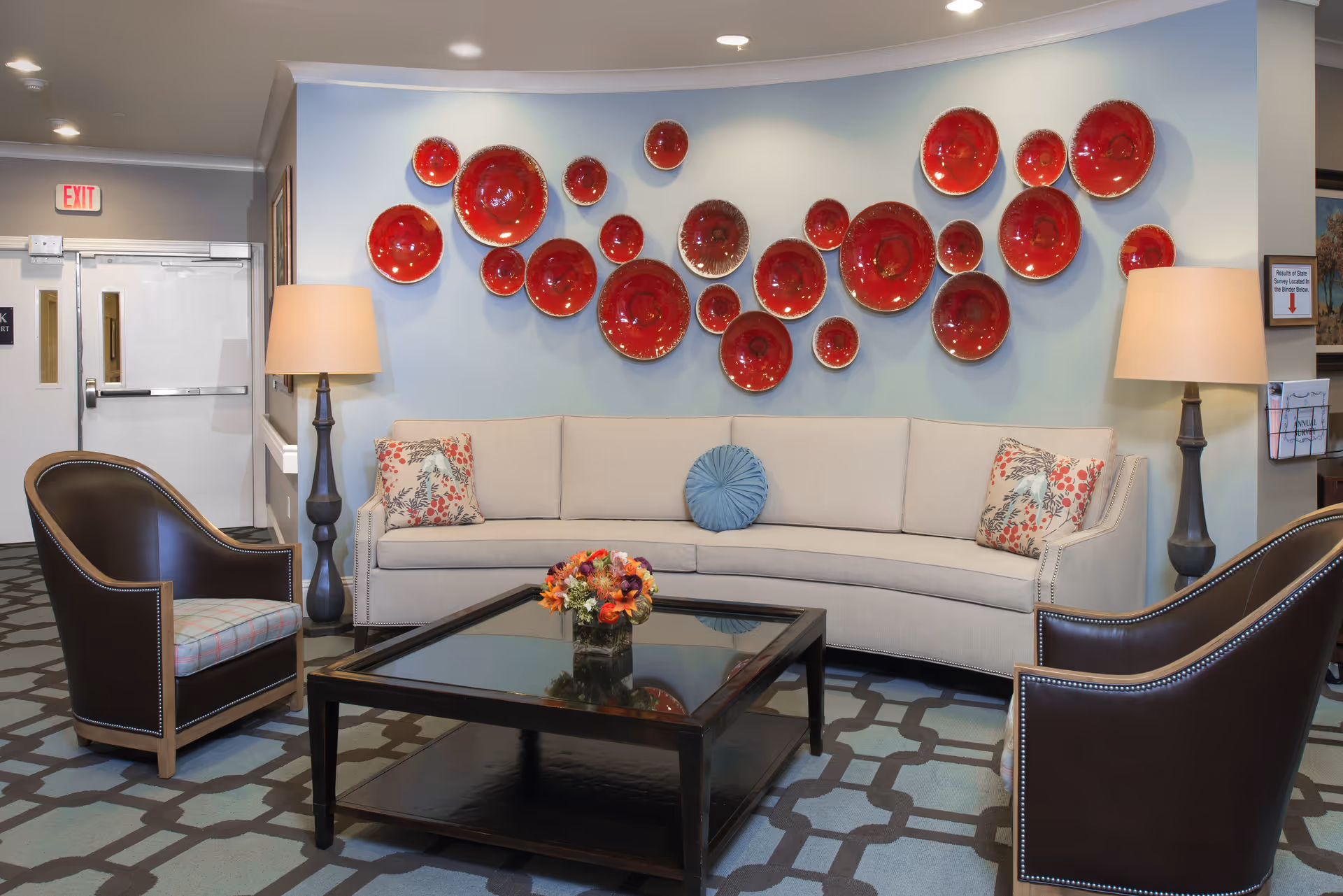 A cozy lobby seating area with a curved beige sofa, two leather armchairs, a glass-top coffee table, lamps, and a wall display of red decorative plates.