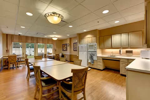 A spacious dining room with wooden floors and multiple tables surrounded by chairs. The room features a kitchen area with beige cabinets, a refrigerator, an oven, a microwave, and a sink. Large windows allow natural light to enter, and ceiling lights provide additional illumination.