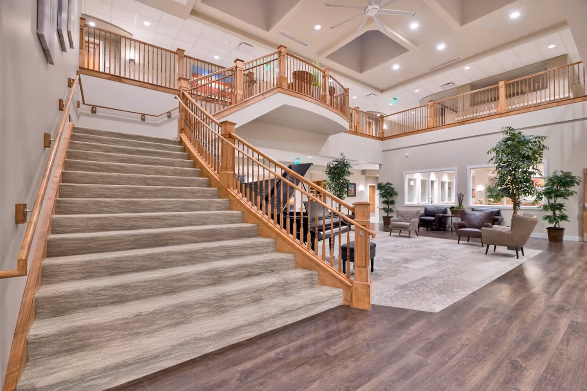 Spacious interior of a senior living facility featuring a wide carpeted staircase with wooden railings leading to an upper level balcony. The area includes a seating arrangement with chairs and sofas, potted plants, and a grand piano. The ceiling is high with recessed lighting and a ceiling fan.